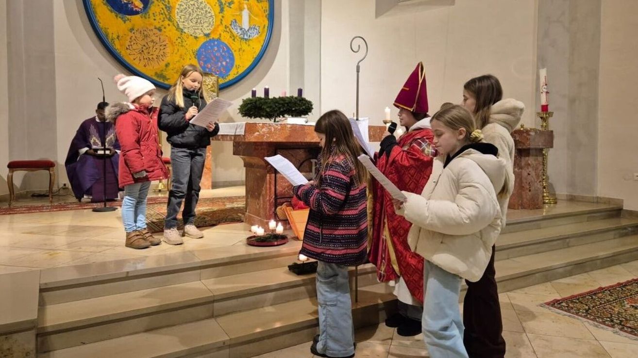 Eine Gruppe von Kindern steht in einer Kirche, hält Papiere in der Hand und liest laut vor während einer Zeremonie. Ein Kind hält ein Mikrofon. Ein Priester in einem roten Gewand steht hinter ihnen.