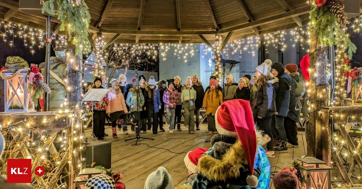Eine Gruppe von Kindern in Winterkleidung steht in einem geschmückten Pavillon und singt mit Musikblättern auf einem Ständer. Sie sind von einem Publikum umgeben.