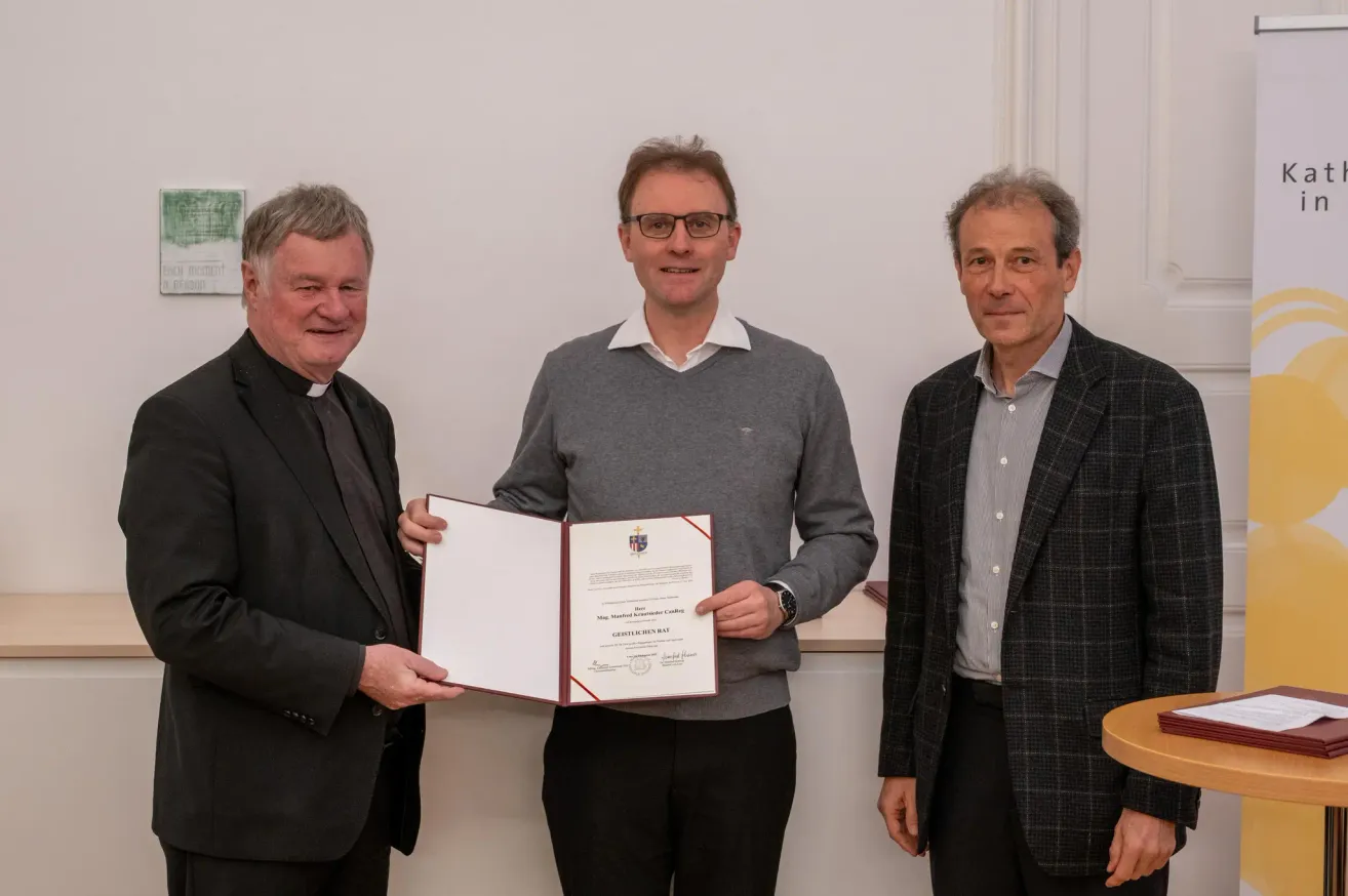 Drei Männer stehen lächelnd vor einer weißen Wand. Der Mann in der Mitte hält ein Zertifikat in der Hand, trägt eine Brille und einen grauen Pullover. Der Mann auf der linken Seite trägt einen schwarzen Anzug und der Mann auf der rechten Seite trägt einen karierten Mantel.