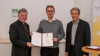 Three men stand in front of a white wall, smiling. The man in the middle holds a certificate, wearing glasses and a gray sweater. The man on the left wears a black suit and the man on the right wears a checkered coat.