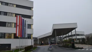 Der Eingang zu einem Gebäudekomplex mit Fahnen auf Stangen und einem großen Vordach. Der Eingang hat ein Stoppschild und eine Schranke. Das Gebäude ist grau und weiß.