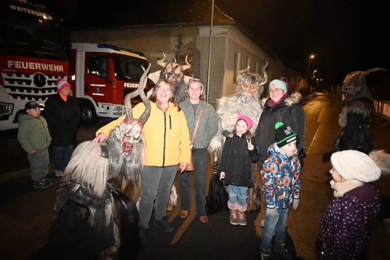 Eine Gruppe von Menschen in Kostümen und Masken posiert für ein Foto auf einer Straße bei Nacht. Eine Person trägt ein Ziegenkostüm. Ein Feuerwehrauto ist im Hintergrund geparkt.