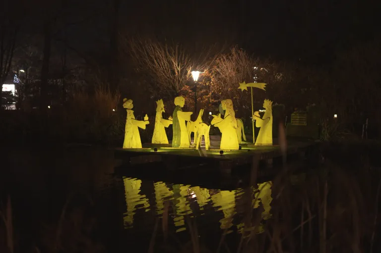 Beleuchtete Figuren der Weihnachtskrippe, einschließlich des Sterns, stehen auf einem Teich mit Reflexionen. Die Szene ist nachts beleuchtet, umgeben von Bäumen und einer Straßenlaterne.