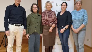 Five people stand in front of a banner with the word 'Familja'. The man wears a black shirt and pants, while the women wear various outfits. They all look in the same direction.