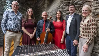 A group of musicians stand behind a hammered dulcimer, with a man holding a cello. Behind them is a stone wall.