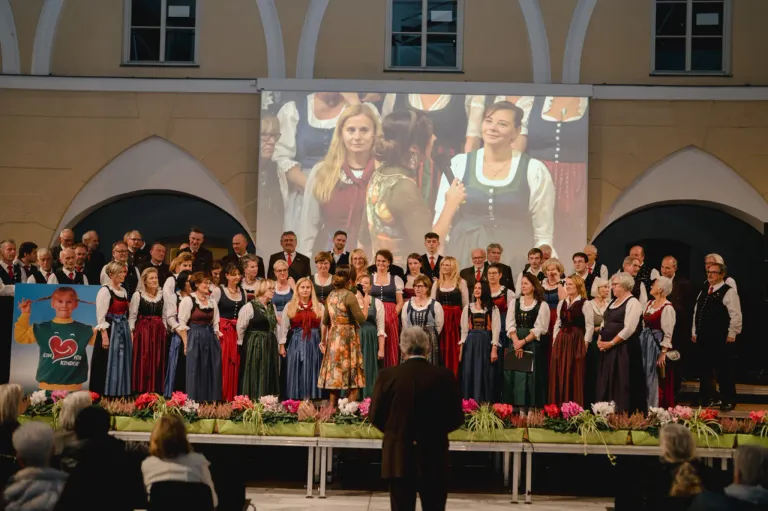 Ein Chor tritt auf der Bühne auf, mit einer großen Leinwand im Hintergrund. Die Chormitglieder sind in traditioneller Kleidung gekleidet. Die Bühne ist mit bunten Blumen geschmückt.