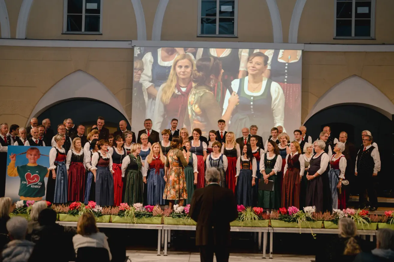 A choir is performing on stage with a large screen behind them. The choir members are dressed in traditional clothing. The stage is adorned with colorful flowers.