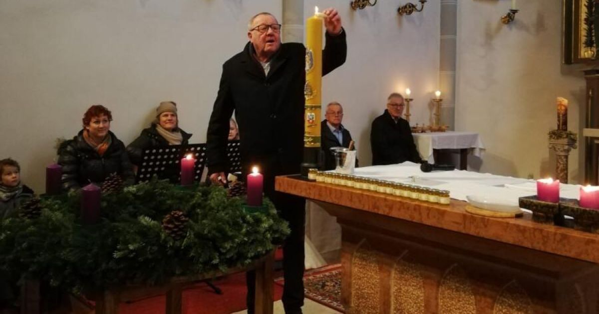 A man stands in a church, holding a large candle with a logo. Candles and a wreath decorate the table. People sit behind him, and a table holds candles and a book.