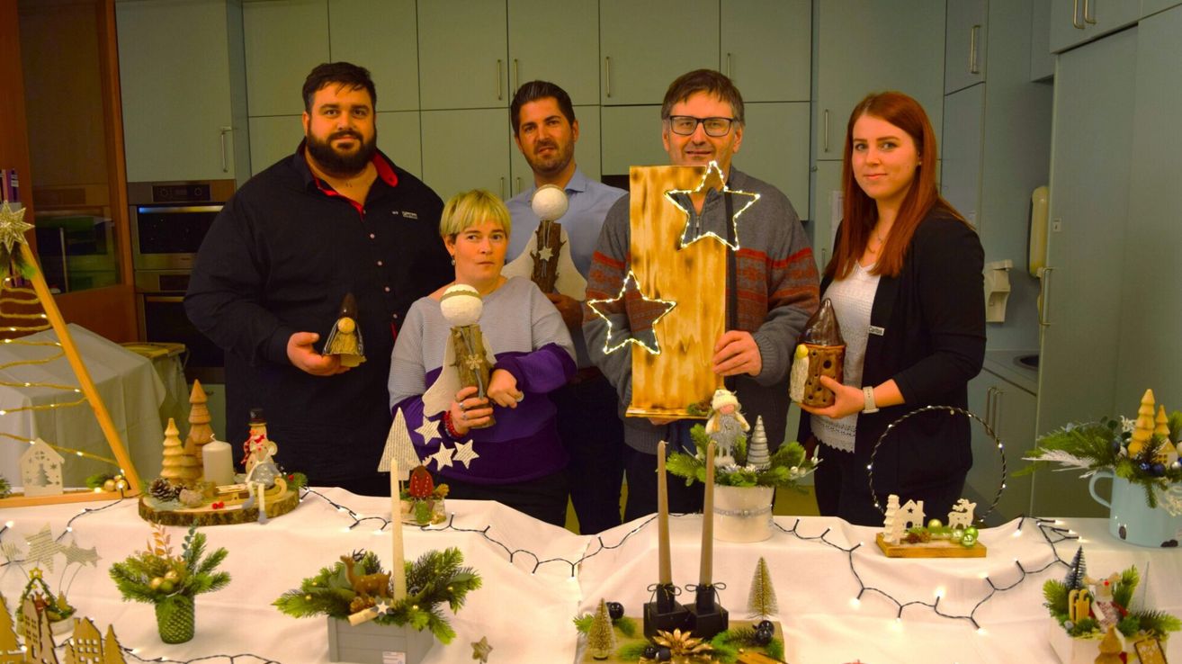 Five people stand behind a table with Christmas decorations. They hold wooden Christmas ornaments. The man on the right holds a wooden star. The man on the left holds a wooden rocket. The woman on the right holds a wooden cup.