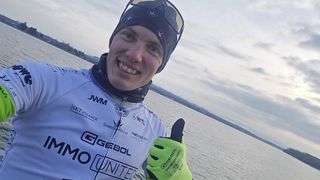 A smiling man in a cycling jersey stands by a body of water, with a seascape in the background. He wears a green and black glove and a cap.