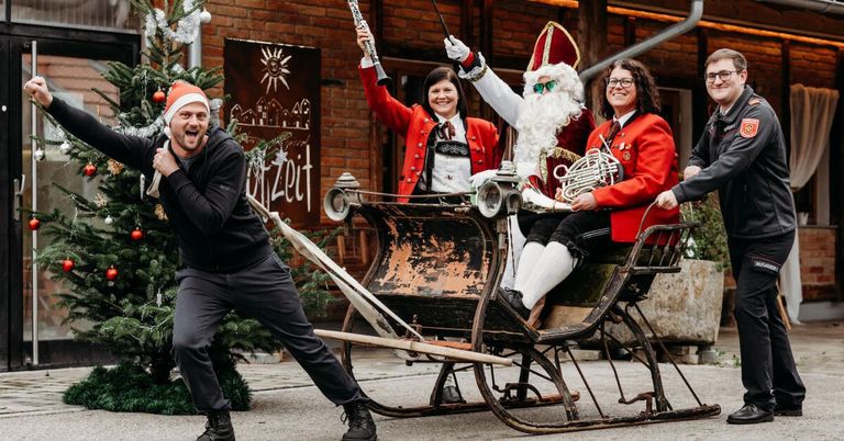 Eine festliche Szene mit dem Weihnachtsmann und seinen Helfern, die in einem Schlitten fahren, wobei eine Person ein Musikinstrument hält.