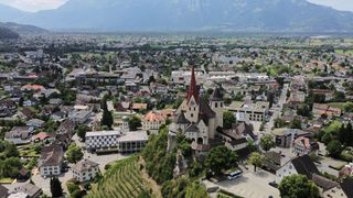 Luftaufnahme einer Stadt mit einer burgartigen Kirche, umgeben von Gebäuden und Bäumen. Berge in der Ferne.
