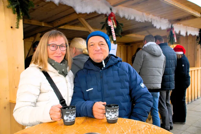 Ein Paar in Winterkleidung sitzt an einem Tisch und hält Kaffeebecher. Sie lächeln, und der Mann trägt eine Mütze. Hinter ihnen stehen zwei Personen, von denen eine eine Jacke mit Kapuze trägt. Über ihnen hängt eine Weihnachtsdekoration.