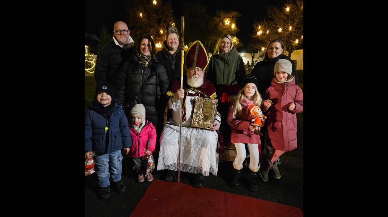 A family and friends pose for a photo with an elderly man dressed as St. Nicholas, holding a book, with children in winter clothes in a festive setting with lit trees.