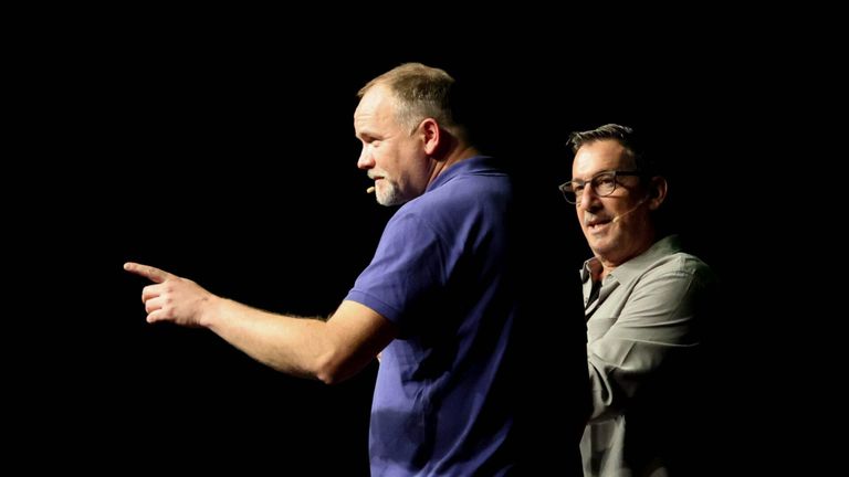 Zwei Männer stehen nebeneinander auf einer Bühne. Der Mann auf der linken Seite hat einen Bart und trägt ein blaues Hemd, während der Mann auf der rechten Seite eine Brille und ein graues Hemd trägt.