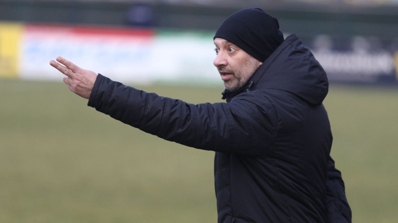 Ein Mann in einem schwarzen Jacke und Mütze zeigt auf einem Fußballfeld mit einem verschwommenen Hintergrund.