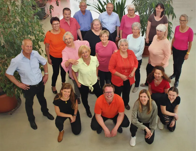 Eine Gruppe von Männern und Frauen, die in verschiedenen Farben gekleidet sind, stehen und hocken zusammen in einem Raum mit Pflanzen.