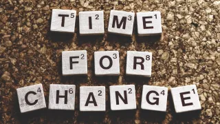 A Scrabble board displaying letters arranged to spell 'TIME FOR CHANGE' on a cork surface. Each letter has a number below it.