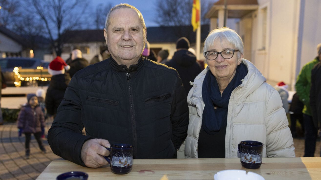 Ein älterer Mann und eine ältere Frau posieren lächelnd für ein Foto, während sie Tassen halten. Sie stehen an einem Tisch mit Tassen und Schalen. Der Hintergrund zeigt ein Gebäude mit einer Fahne und Leuten.