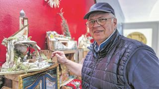 An older man wearing glasses and a hat stands next to a cabinet with a Christmas scene. The background features a red wall.