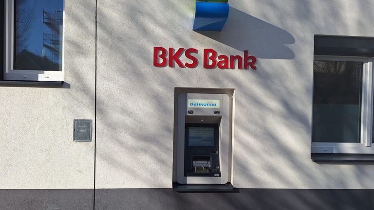 Ein Geldautomat der BKS Bank ist an der Wand montiert. Es ist ein weißer Automat mit einem blauen Schild mit der Aufschrift 'Bankomat' darüber.