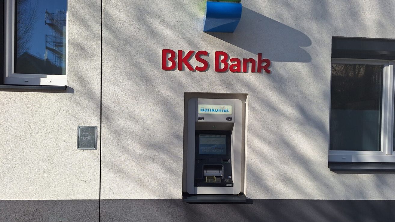 Ein Geldautomat der BKS Bank ist an der Wand montiert. Es ist ein weißer Automat mit einem blauen Schild mit der Aufschrift 'Bankomat' darüber.