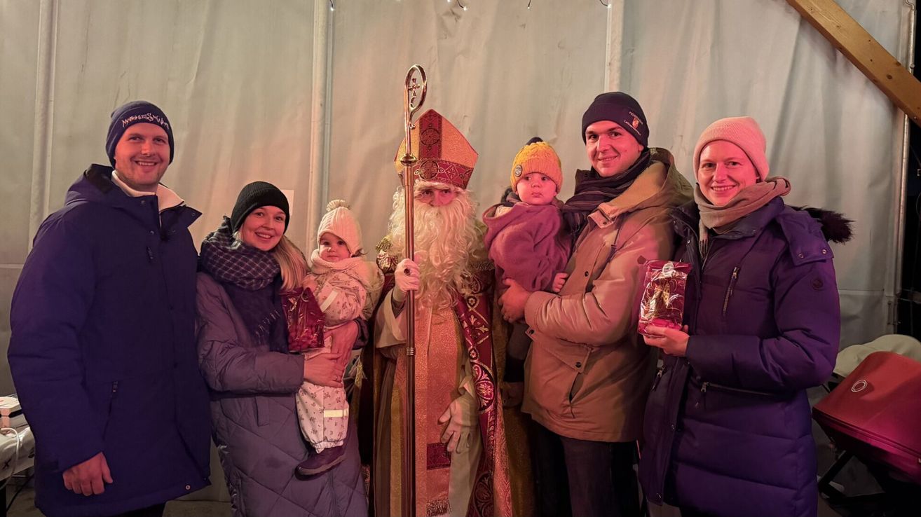 Eine Familie posiert mit einem Mann, der als Sankt Nikolaus gekleidet ist. Sie alle tragen Winterkleidung und lächeln. Zwei Babys sind im Bild, eines wird von einer Frau und das andere von einem Mann gehalten.