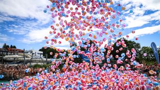 A colorful cloud of beach balls floats in the air above a crowd of people on a sunny day. Trees and a building are in the background.