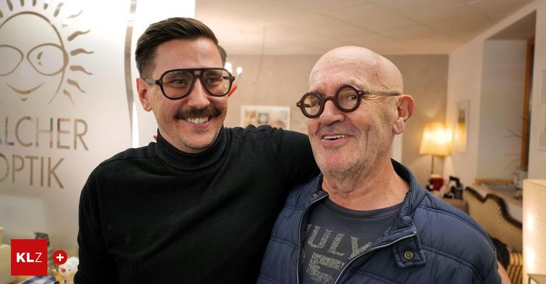 Two men are smiling in a room, one with glasses and the other with a mustache, standing close to each other.