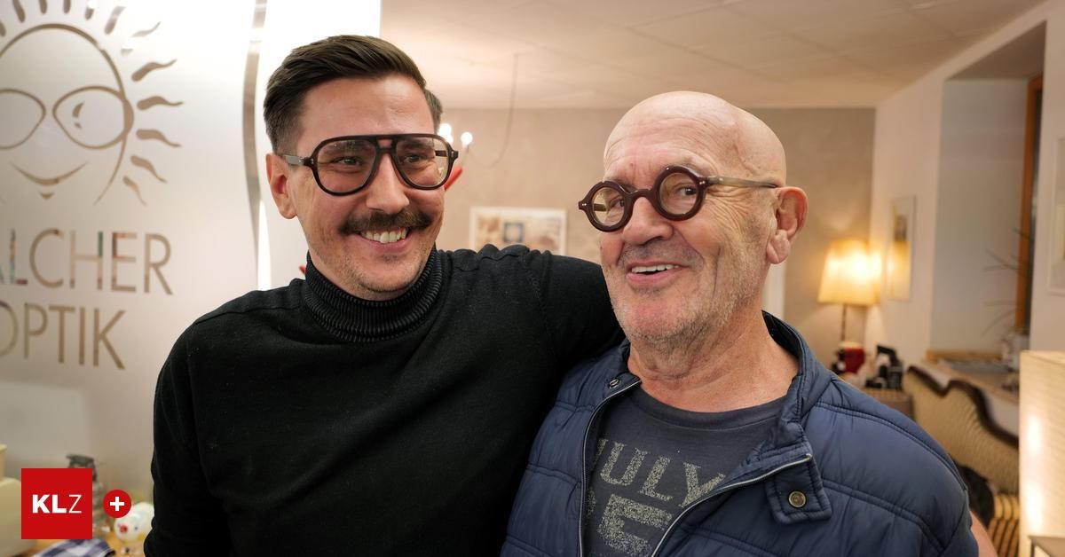 Two men are smiling in a room, one with glasses and the other with a mustache, standing close to each other.