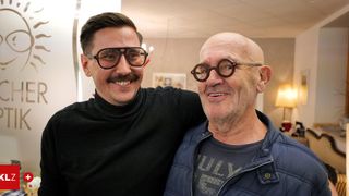 Two men are smiling in a room, one with glasses and the other with a mustache, standing close to each other.
