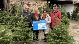 Four people stand outside, posing for a photo, with a donation check for 1000 Euros. The check is for the Schwarzeitaler Social Club. Behind them, there are pine trees and a brick building with solar panels.
