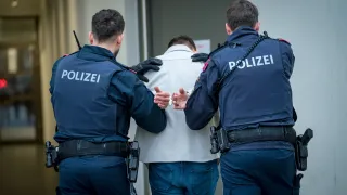 Three police officers are escorting a person in a white jacket and jeans. The person is handcuffed and being led through a door.