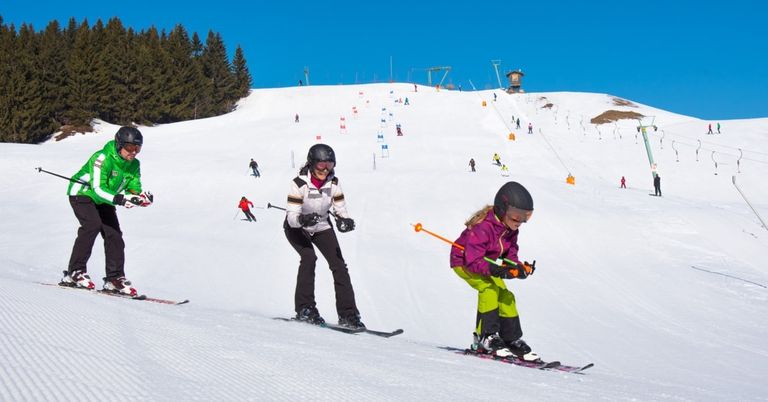 Skifahrer fahren auf einer verschneiten Piste. Eine Frau und ein Mädchen fahren gemeinsam. Das Mädchen trägt eine Schutzbrille und hält Skistöcke. Bäume und Berge sind im Hintergrund.