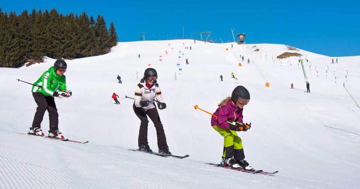 Skifahrer fahren auf einer verschneiten Piste. Eine Frau und ein Mädchen fahren gemeinsam. Das Mädchen trägt eine Schutzbrille und hält Skistöcke. Bäume und Berge sind im Hintergrund.