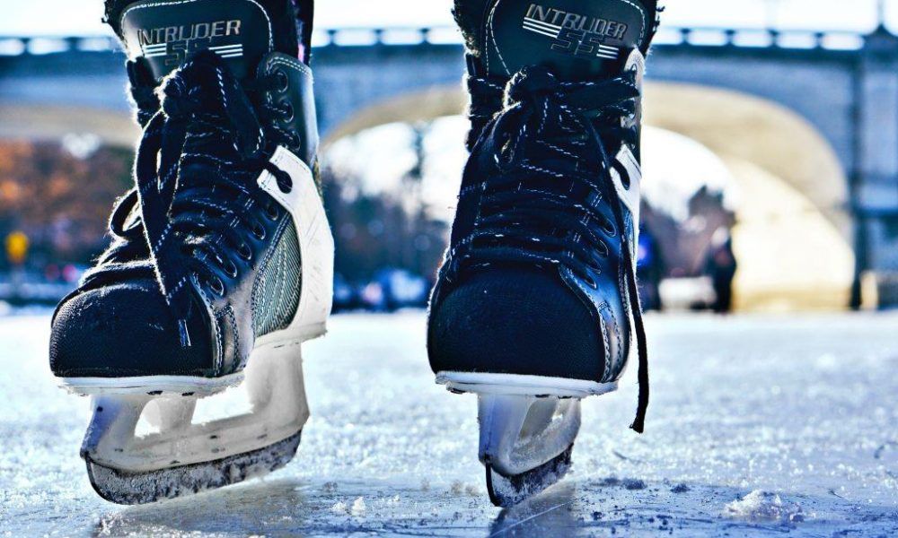 A close-up view of black hockey skates with white trim, blades, and the brand name 'Intruder' is visible on the ice. The background shows a blurry bridge.