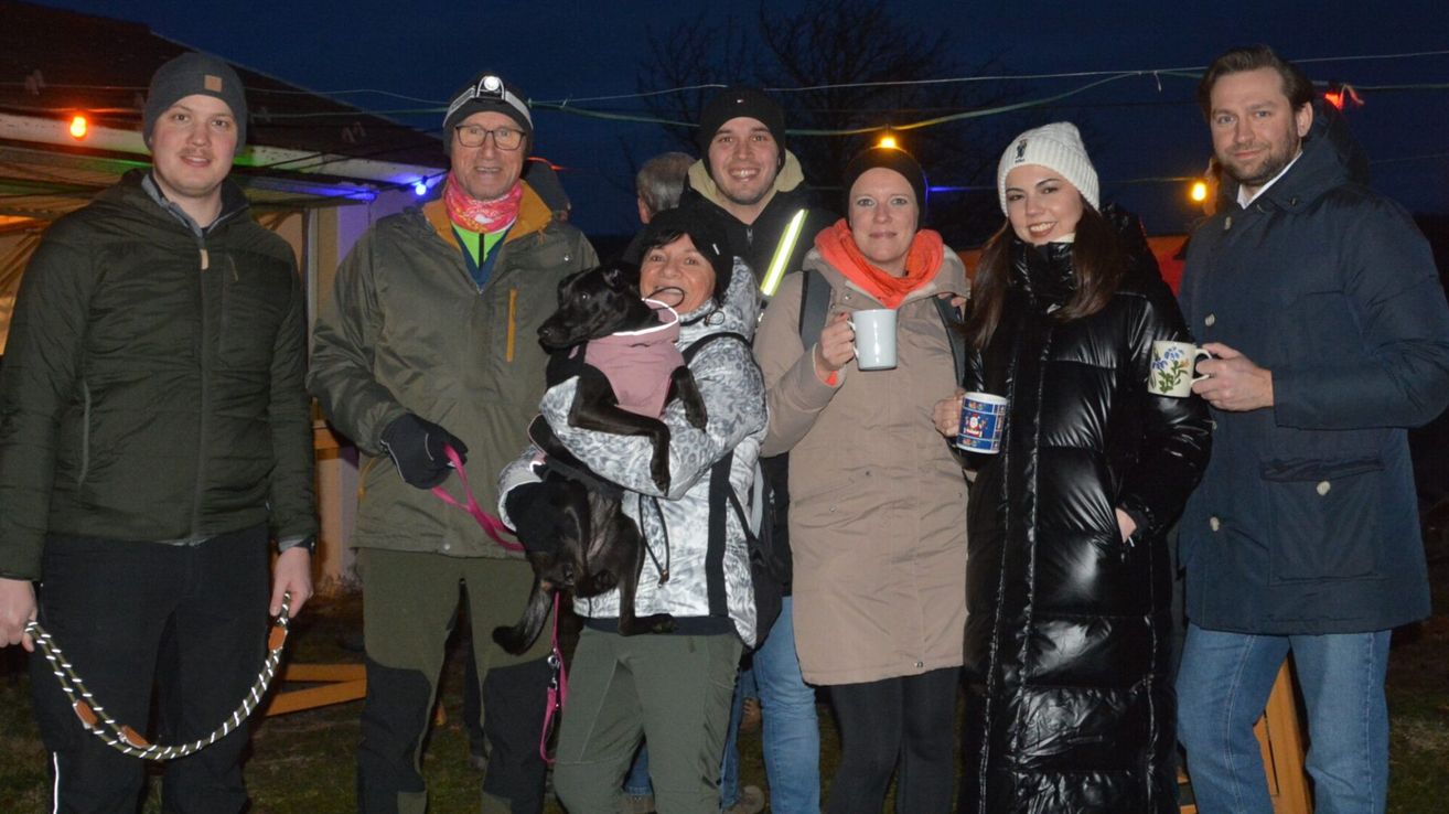 Fünf Personen stehen in einer Gruppe mit einem schwarzen Hund in Winterkleidung bei Nacht. Der Mann links hält eine Leine. Eine Frau in der Mitte hält einen Becher. Eine Frau rechts lächelt. Ein Mann hinter ihnen trägt eine Warnweste. Bäume und Lichter sind im Hintergrund.