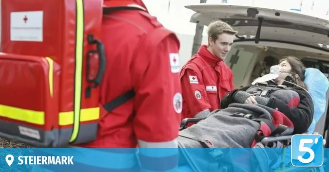A woman lies on a stretcher in an ambulance. Two men in red uniforms assist her. The ambulance is open, and the woman has an oxygen tube in her nose. The ambulance has a logo with the number 5.