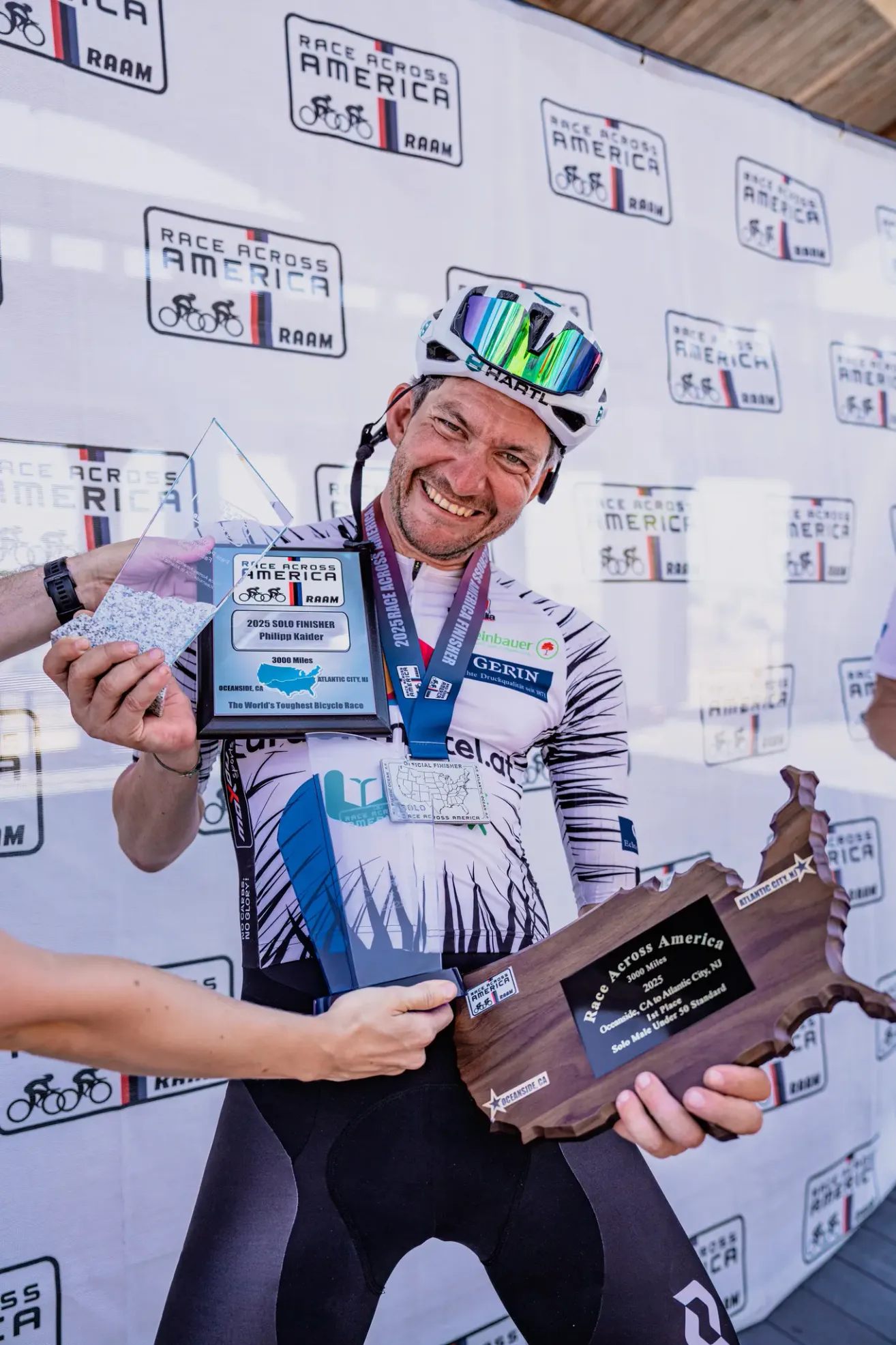 A cyclist poses with a trophy and medals, wearing a helmet and cycling gear. The backdrop is a wall with logos and text related to a bicycle race.