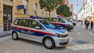 Two police cars are parked on a street in front of a building with a sign reading 'POLIZEI'. The cars have 'POLIZEI' written on them, and there are people walking in the background.