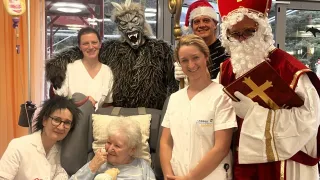 A group of people dressed in festive costumes, including two as Santa Claus and a werewolf, are posing for a photo with an elderly woman in a hospital bed.