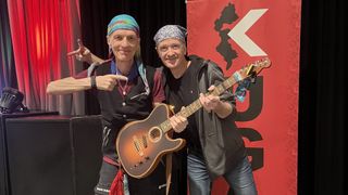 Two men stand together, one holding a guitar, wearing bandanas and smiling, in front of a red banner.