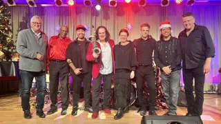 A group of musicians in various attires stand on a stage, likely for a Christmas celebration. One man holds a saxophone. Behind them is a Christmas tree with ornaments and spotlights.