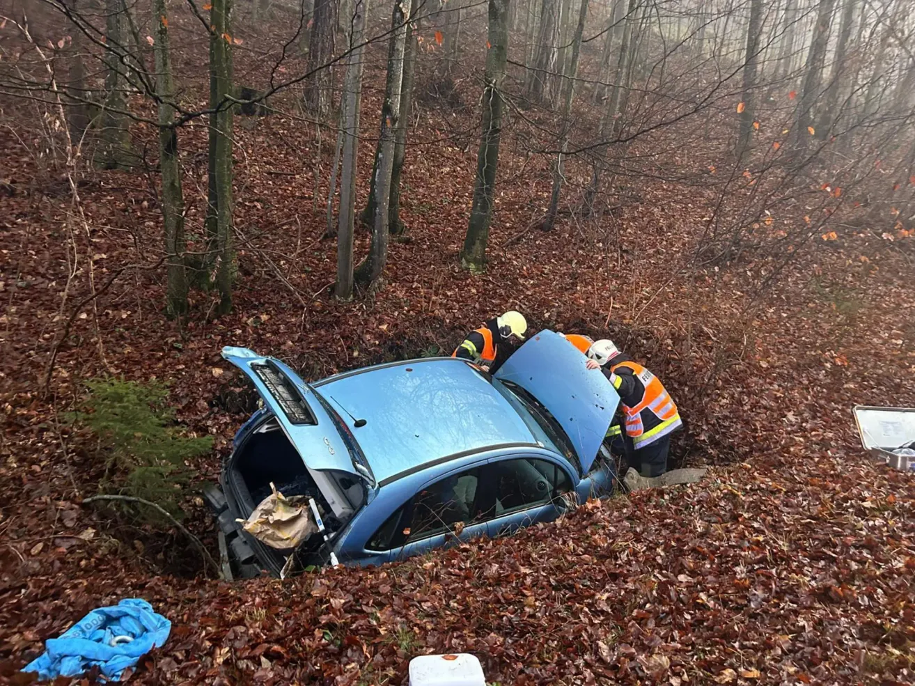 Ein blaues Auto ist in einem Wald umgekippt, wobei zwei Personen in Warnwesten daran arbeiten. Der Kofferraum des Autos ist offen und Trümmer sind umher verstreut.