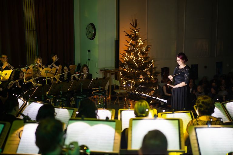 Eine Gruppe von Musikern tritt vor einem geschmückten Weihnachtsbaum und einer Wanduhr auf. Die Dirigentin hält einen Taktstock und lächelt, während das Publikum aufmerksam zuhört.
