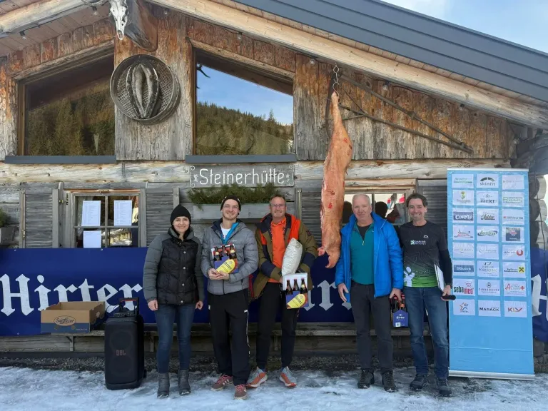 Fünf Personen posieren für ein Foto vor einer Skihütte im Schnee. Sie halten Flaschen Bier und ein großes Stück Fleisch. Ein Schild mit dem Text 'Steinerhütte' befindet sich über ihnen.