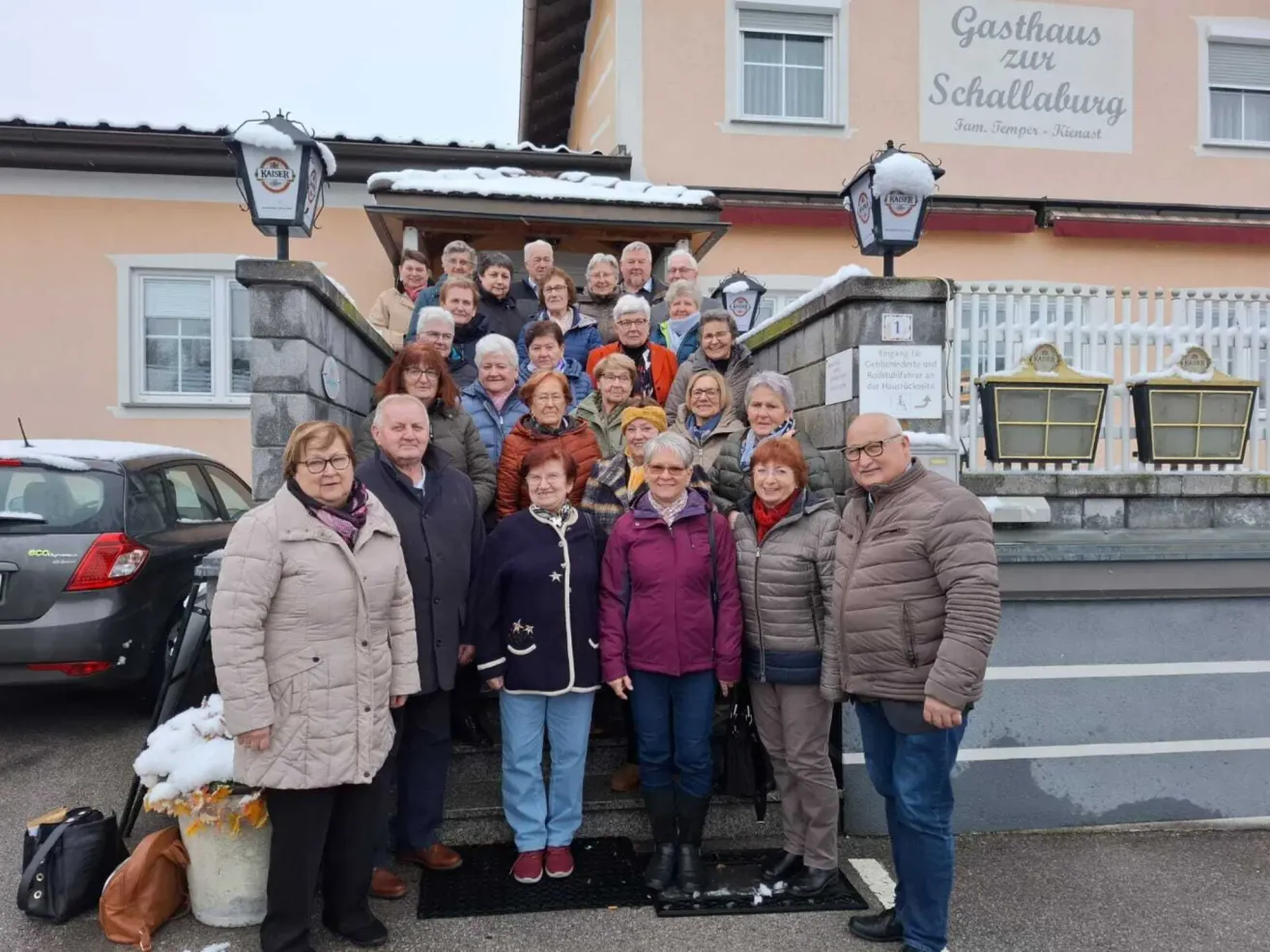 Eine Gruppe von Menschen steht vor einem Gebäude mit Schnee auf dem Dach. Sie tragen alle Winterkleidung und einige lächeln. Das Gebäude hat ein Schild mit der Aufschrift 'Gasthaus zur Schallburg'.