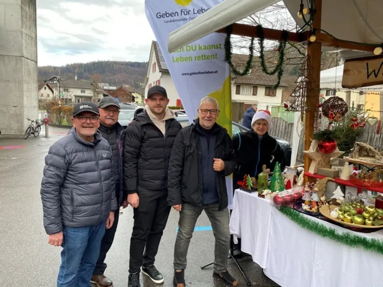 Fünf Männer stehen lächelnd zusammen vor einem Weihnachtsmarktstand. Hinter ihnen steht ein Tisch, der mit verschiedenen Weihnachtsdekorationen beladen ist. Ein Schild mit deutschem Text ist sichtbar.