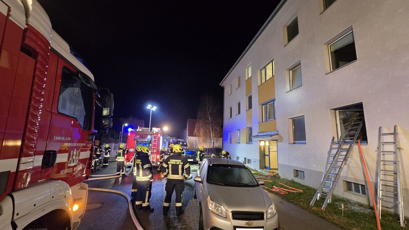 Firefighters stand near a building at night, with one of them holding a hose. A fire truck is parked nearby. The scene is illuminated by street lights and car headlights.