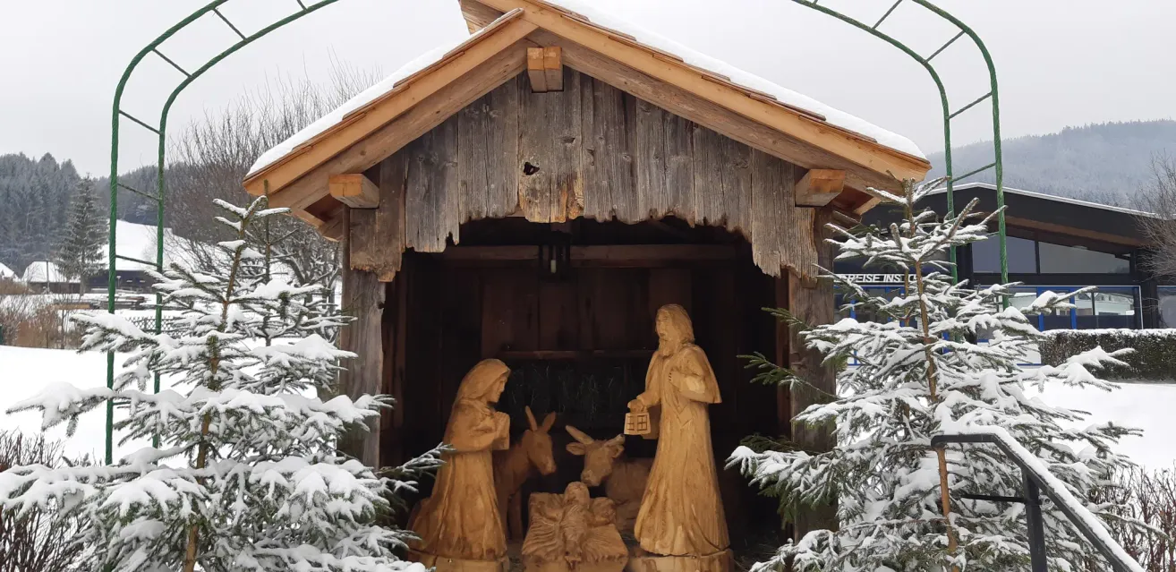 Eine hölzerne Krippe in einer rustikalen Scheune ist mit Schnee bedeckt. Sie zeigt Maria, Josef und das Jesuskind mit einem Esel und einem Schaf.